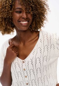 Biała sweter z dzianiny z dekoltem w kształcie litery V i krótkimi rękawami, z charakterystycznym wzorem w zygzaki oraz trzema drewnianymi guzikami z przodu.