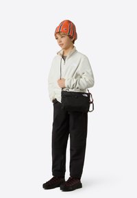 Czarna torebka na ramię z karabińczykiem, noszona przez dziecko w białym polarze, pomarańczowej czapce z dzianiny w paski, czarnych spodniach i ciemnych butach.