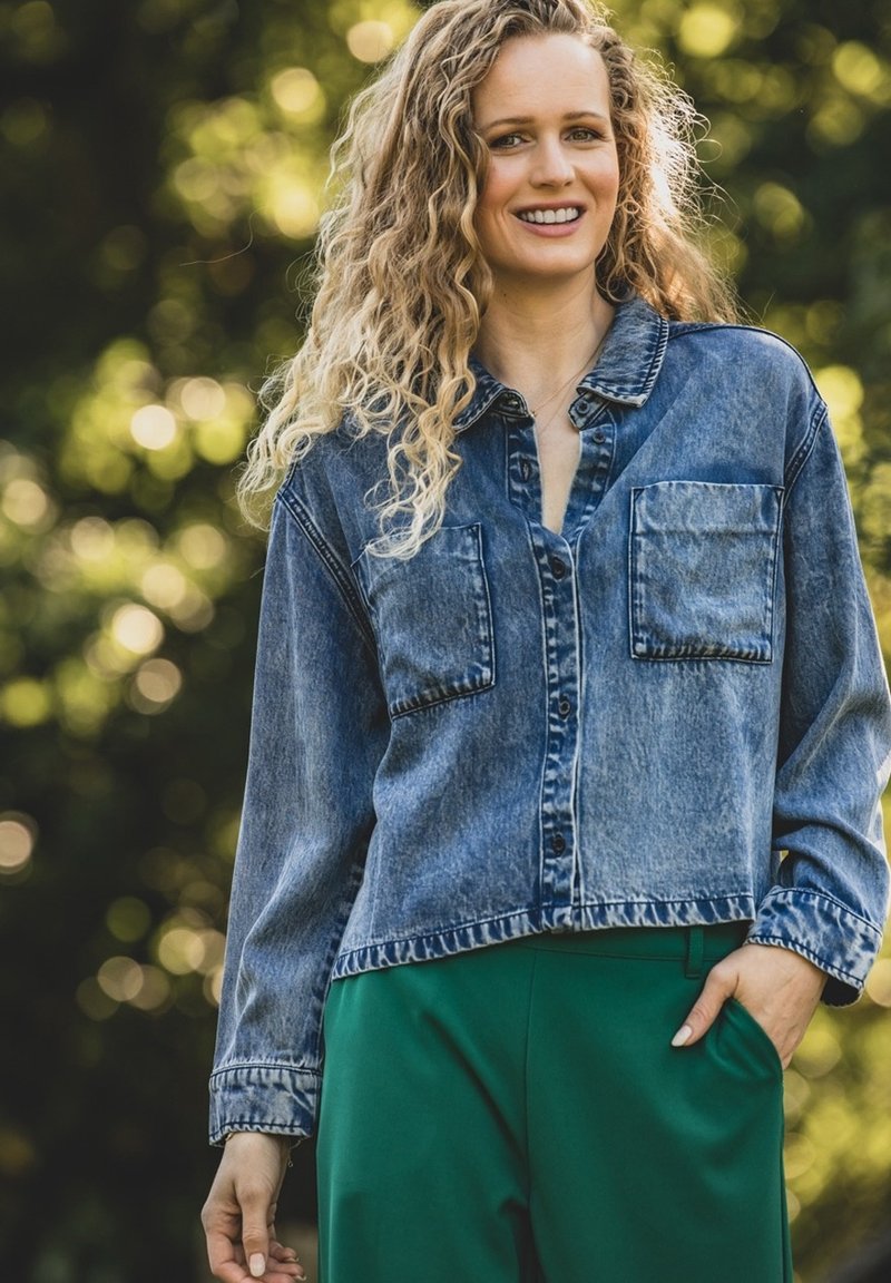 Denimjack met een cropped ontwerp, knoopsluiting aan de voorkant, twee borstzakken en een vervaagde blauwe kleur. Gecombineerd met groene broek.