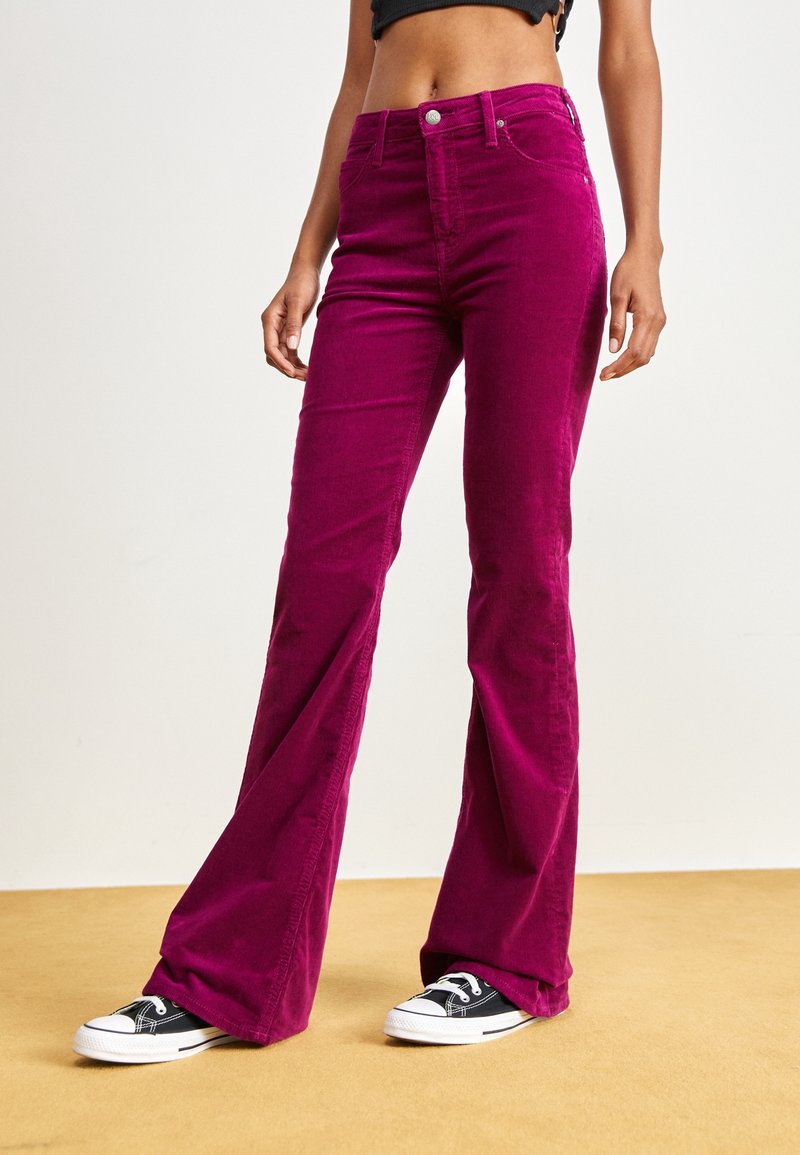Femme portant un pantalon flare en velours côtelé magenta taille haute et des baskets basses noires, debout sur un sol beige avec un fond blanc uni.