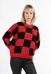 Rood en zwart geblokt sweater met een textured garen, met lange mouwen en een geribde halslijn. Draagt over strakke zwarte broeken.