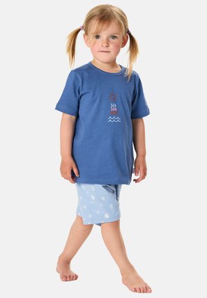 Junges Mädchen mit blonden Zöpfen, das ein blaues "Sea Sun Sand" T-Shirt und hellblaue gemusterte Shorts trägt, barfuß auf weißem Hintergrund läuft.
