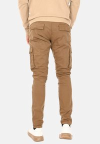 Pantaloni cargo khaki con due tasche laterali, vestibilità slim, realizzati in tessuto misto cotone, caratterizzati da una leggera lucentezza e polsini alla caviglia, abbinati a sneaker bianche.