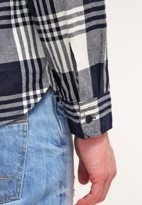 Marineblauw en witte geruite blouse met lange mouwen en knoopmanchetten, gemaakt van zacht stof. Draagt met lichtblauwe denim jeans.