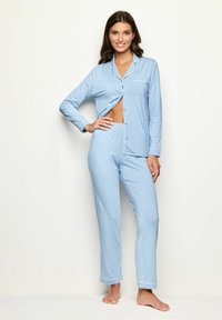 Lichtblauw pyjama set met een knoopjes top en een zak, en lange broek. Gemaakt van zachte stof met witte piping langs de randen.