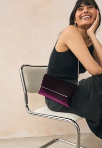 Sac à épaule violet en velours de forme rectangulaire, avec des garnitures en argent et une fine bandoulière, posé sur les genoux d'une personne assise.