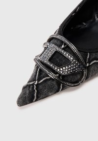 Μαύρο υφασμάτινο παπούτσι με μυτερή μύτη και λευκές ραφές, διακοσμημένο με μια μεγάλη αγκράφα στολισμένη με στρας σε ασημί τόνο στη μύτη.