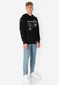 Jonge man die staat in een zwarte hoodie met witte technische grafische afbeeldingen, lichtblauwe spijkerbroek en witte sneakers tegen een witte achtergrond.