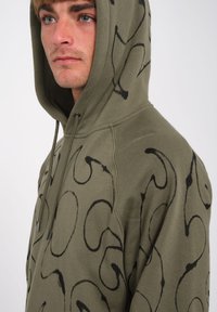 Felpa con cappuccio verde oliva realizzata in tessuto morbido con un motivo astratto nero, dotata di tasca frontale e cappuccio regolabile con cordino.
