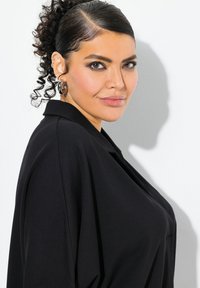 Černá oversized blůza s výstřihem do V a hladkým povrchem, s krátkými rukávy. Modelka má kudrnaté vlasy a kruhové náušnice.