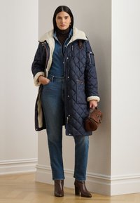 Καπιτονέ μπλε σκούρο παλτό με εκρού γιακά από shearling, καφέ δερμάτινες λεπτομέρειες και τσέπη με φερμουάρ. Το μοντέλο φορά τζιν και καφέ μπότες με τακούνι.