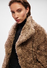 Καφέ φανελένιο μπουφάν με μεγάλο γιακά, κατασκευασμένο από υφασμένο ύφασμα. Φοριέται πάνω από ένα μαύρο φανελάκι με ψηλό λαιμό, διαθέτοντας ορατά κουμπιά και χαλαρή εφαρμογή.