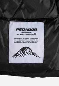 Svart ytterstoff med et hvitt merke. Merket har et fjellgrafikk, merkevarenavnet "Pegador" og tekst om eventyrlig bruk.