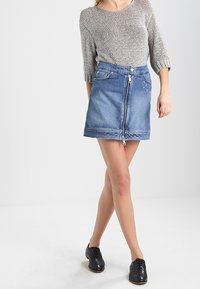 Hame denim dengan ritsleting depan, warna biru, desain lima saku, dan hem yang kontras. Dipadukan dengan atasan rajut lengan pendek berwarna abu-abu dan sepatu hitam.