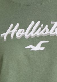 Olivgrön sweatshirt i mjukt material med vit blockbokstavstext "Hollister" och en stiliserad fågallogo nedanför.
