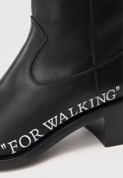 Crne kožne čizme do gležnja s izduženim vrhom, s bijelim natpisom "ZA HODANJE" uz bočnu stranu i niskom blok petom.
