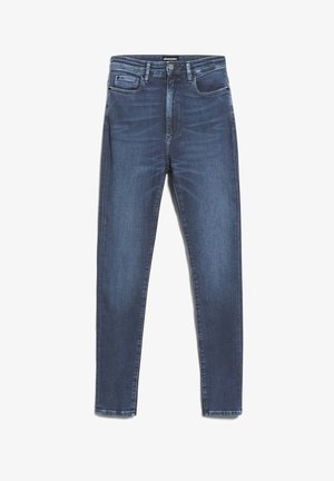 Kobieta w niebieskich dopasowanych dżinsach z wysokim stanem, białej koszulce z krótkim rękawem i dużych beżowych butach typu slip-on, stoi na białym tle.