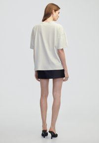Světle šedé oversized tričko s krátkými rukávy, prezentované na modelce nosící černou mini sukni a mules na blokovém podpatku, ukazující hladkou texturu.