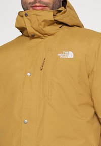 Man som bär en senapsgul The North Face-jacka med hög krage, blixtlåsförsedd bröstficka och märkta tryckknappar framtill.