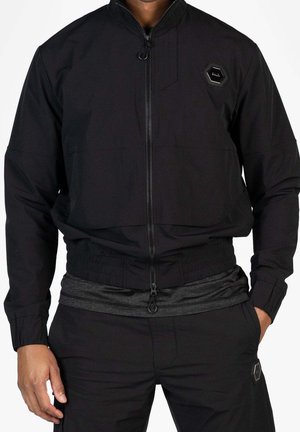 Mann trägt schwarze Reißverschlussjacke mit elastischem Bund, passende schwarze Hose und dunkelgraues Shirt darunter, eine Hand in der Tasche.