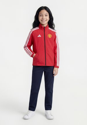 Pige står og smiler iført en rød Manchester United jakke med hvide striber, marineblå bukser og hvide sneakers mod en hvid baggrund.