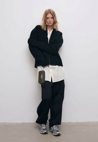 Svart stickad oversized jacka över en vit skjorta, tillsammans med lösa svarta byxor och grå sneakers. En grön påse hänger från jackan.