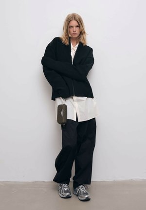 Sort strikket oversized jakke over en hvid skjorte, parret med løstsiddende sorte bukser og grå sneakers. En grøn taske hænger fra jakken.