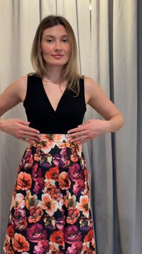 Femme aux cheveux blonds lisses portant un haut noir sans manches et une jupe fleurie, ajustant la taille, debout devant un fond de rideau uni.