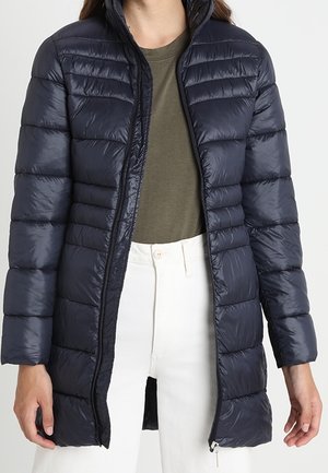 Vrouw draagt een lange, zwarte donsjas over een olijfgroen shirt en witte broek met hoge taille, met de jas opengeritst.