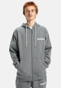 Grijze rits hoodie gemaakt van zacht materiaal. Heeft een trekkoordkap, twee voorzakken en een logo in het wit gedrukt op de borst.