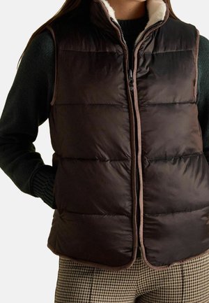 Kvinde iført en mørkebrun polstret vest med beige kant, sort sweater og brune hundetandsbukser, hænder i vestens lommer.
