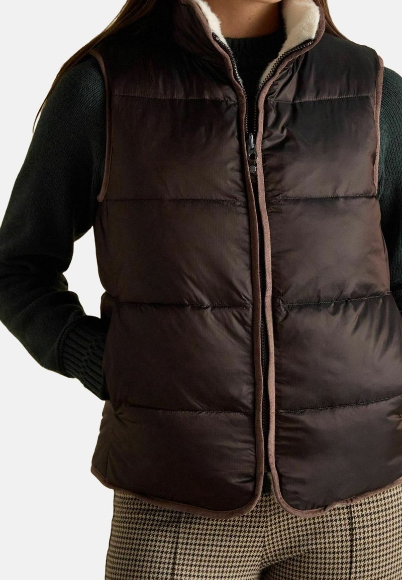 Femme portant un gilet matelassé brun foncé avec un bord beige, un pull noir et un pantalon marron à motif pied-de-poule, mains dans les poches du gilet.