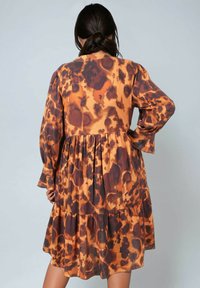 Femme aux cheveux foncés attachés, portant une robe ample à manches longues avec un imprimé animal orange et marron, se tenant avec une main sur la hanche.