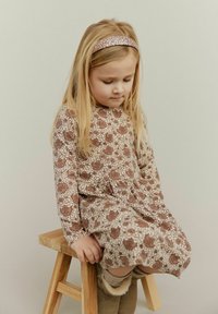 Smeđa cvjetna haljina s dugim rukavima, strukirana u struku, s okruglim izrezom. Uparena s bež čizmama i sjajnim trakom za kosu. Svijetla pozadina.