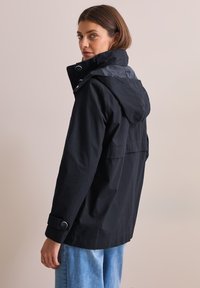 Svart vattentät jacka med huva, texturerad tyg, knäppta manschetter och en ryggdesign med ok. Modell bär ljusblå jeans.