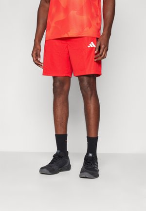 Red athletic shorts with a mesh texture and side pockets, paired with black sports shoes and black ankle socks. Adidas logo on shorts.