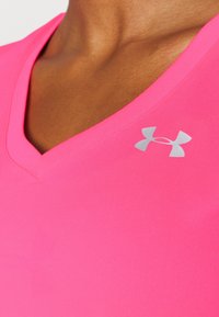 Světle růžové tričko s výstřihem do V vyrobené z elastického materiálu, na levé části hrudi s malým stříbrným logem Under Armour. Hladký povrch.