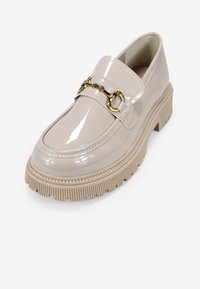 Zapato estilo mocasín de charol color beige con suela gruesa, que presenta un detalle de cadena dorada y un diseño de punta redonda.