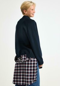 Mörkblå tröja med rund halsringning och ribbade muddar, lager-på-lager över en röd- och vitrutig skjorta som syns vid fållen. Blå denimjeans.