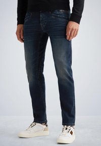 Vaqueros azul oscuro de corte slim, con bolsillos delanteros y traseros, desvanecimiento sutil, y complementados con zapatillas blancas con acentos beiges.