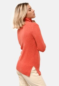 Maglione a collo alto color corallo realizzato in morbido tessuto a maglia, con dettagli a coste e spacchetti laterali. Indossato con pantaloni chiari.