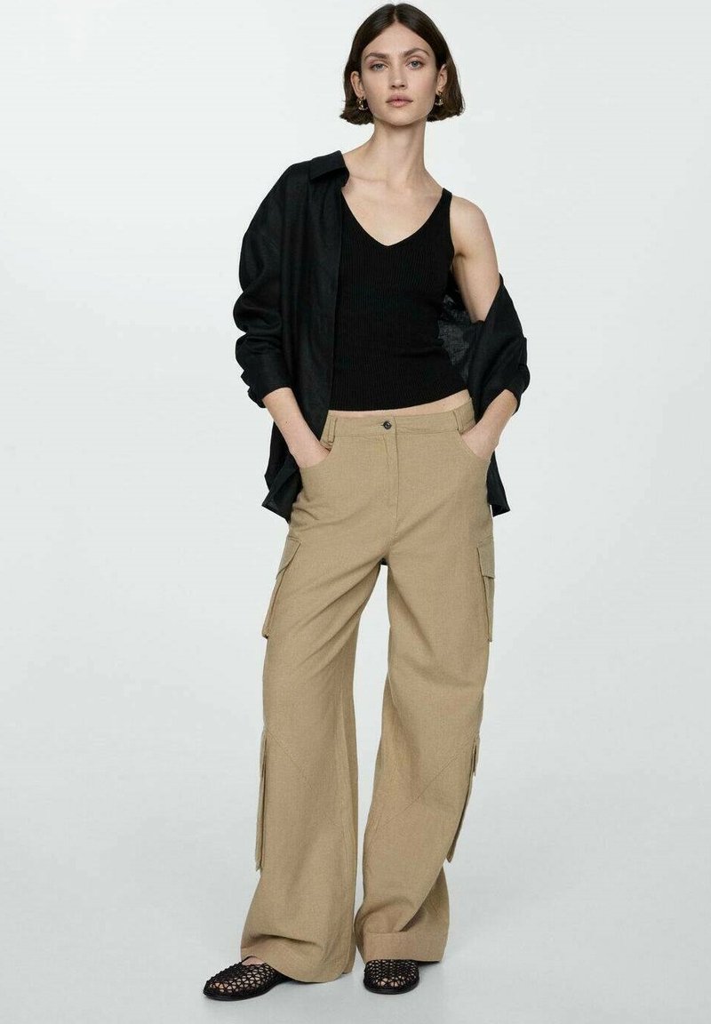 Beżowe spodnie cargo o szerokich nogawkach, z bocznymi kieszeniami i wysokim stanem, połączone z czarnym topem bez rękawów oraz otwartą czarną koszulą. Czarne buty wsuwane.