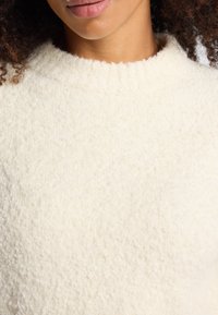 Maglione color crema con una superficie morbida e texture, caratterizzato da un colletto rotondo e bordi a coste. Nessun motivo o accento visibile.