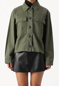 Olivově zelená zkrácená džínová bunda s límcem, zapínáním na knoflíky vpředu a dvěma náprsními kapsami. Nasažená přes černou koženou sukni.