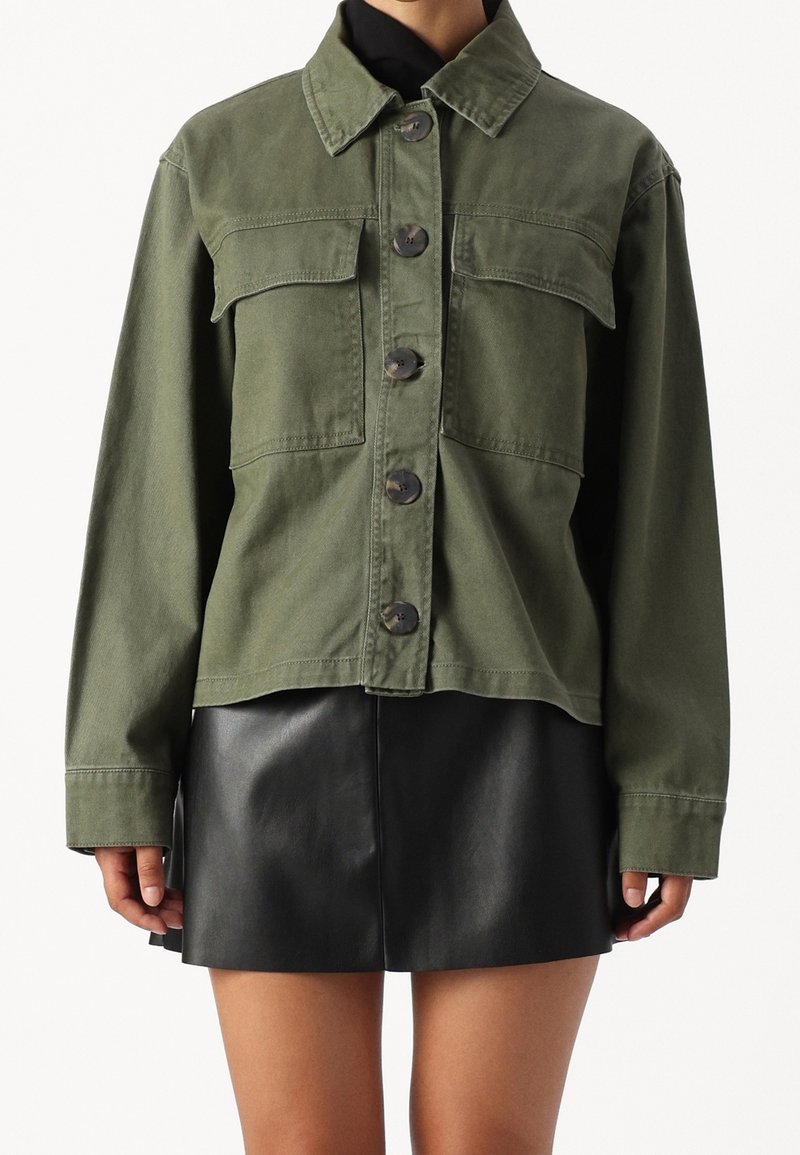 Olivově zelená zkrácená džínová bunda s límcem, zapínáním na knoflíky vpředu a dvěma náprsními kapsami. Nasažená přes černou koženou sukni.
