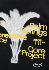 Zwarte t-shirt met witte en gele grafische tekst en een palmblaadje ontwerp, met "Palm Springs" en "Core Project" prominent weergegeven.