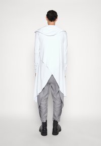 Lunga felpa bianca con cappuccio e orlo asimmetrico, abbinata a pantaloni grigi chiari a sigaretta e stivaletti neri alla caviglia, vista da dietro.