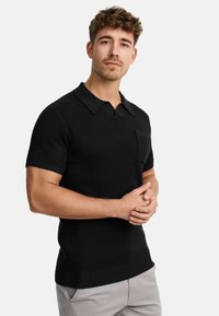 Crna kratka majica polo s rebrastom teksturom, ravnim ovratnikom i džepom na grudima. Model stoji s prekriženim rukama, odjeven u svijetlosive hlače.