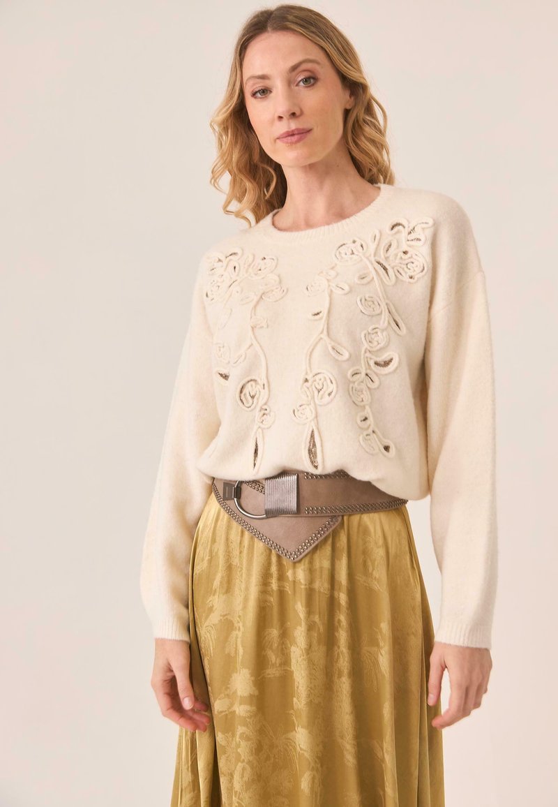 Suéter de punto crema con bordado floral, combinado con una falda fluida de color verde oliva que presenta detalles texturizados y un cinturón ancho beige.