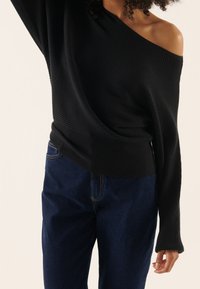 Femme portant un pull côtelé noir à épaules dénudées et un jean bleu foncé, un bras levé et des cheveux bouclés visibles.